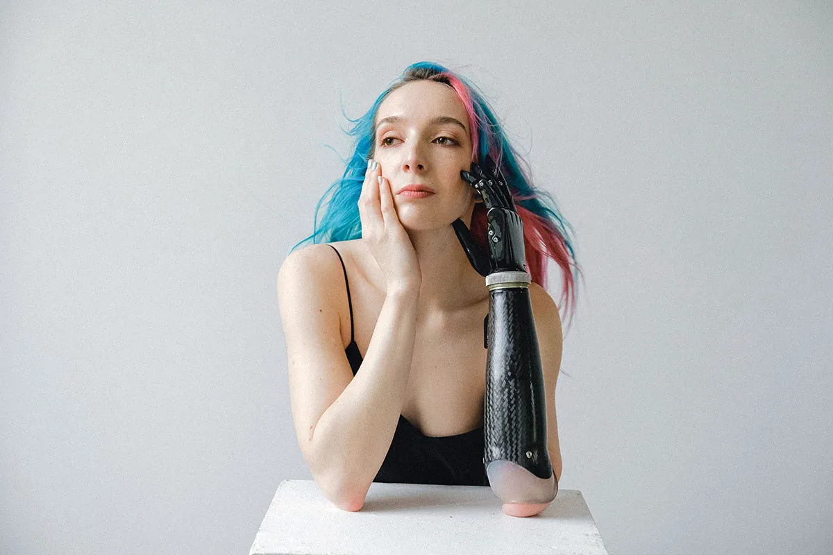 Portrait. Frau mit pink-blauen Haaren stützt Kopf auf Händen ab und blickt in die Ferne